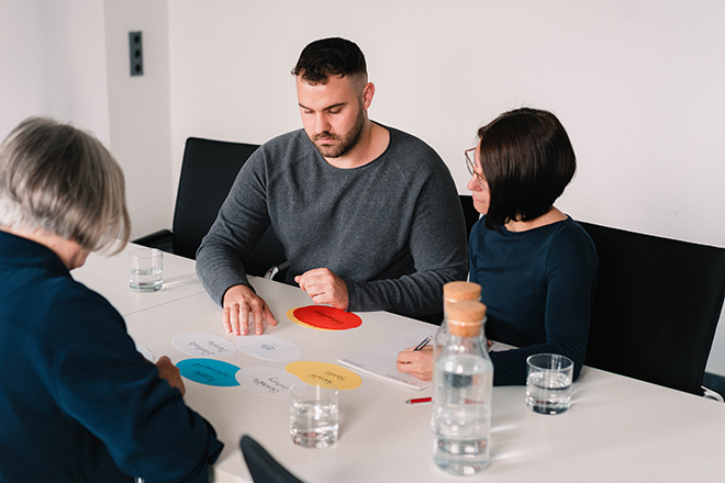 Zwei Frauen und ein Mann sitzen an einem Tisch und besprechen farbige Konzeptkarten während eines Workshops;